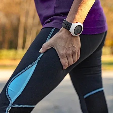 A person outdoors grasps the upper leg during activity, wearing athletic clothing and a fitness watch.